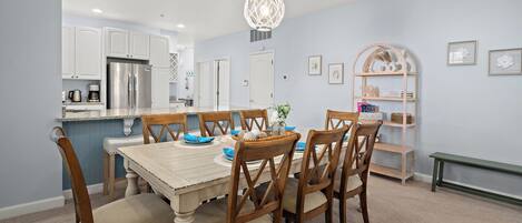 A dining area with a wooden table and chairs, a chandelier, and a kitchen in the background.