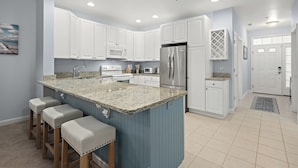 A modern kitchen with white cabinets, a blue island with stools, stainless steel appliances, and a tiled floor.