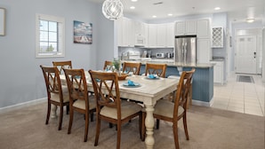 A dining area with a wooden table and chairs, a chandelier, and a kitchen with white cabinets and stainless steel appliances.