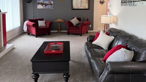 Ein Wohnzimmer mit einem schwarzen Ledersofa, roten Sessel, einem schwarzen Couchtisch und einem roten Beistelltisch. An der Wand hängt eine Uhr, eine gerahmte Karte und eine Lampe.