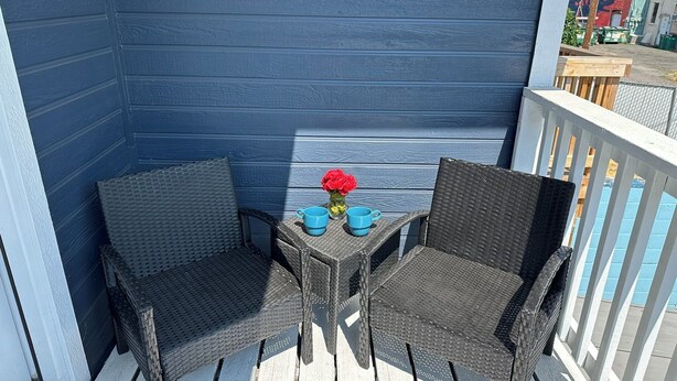 Ein kleiner Balkon mit zwei Korbstühlen, einem Tisch, einer Vase mit roten Blumen und einem blauen Geländer.
