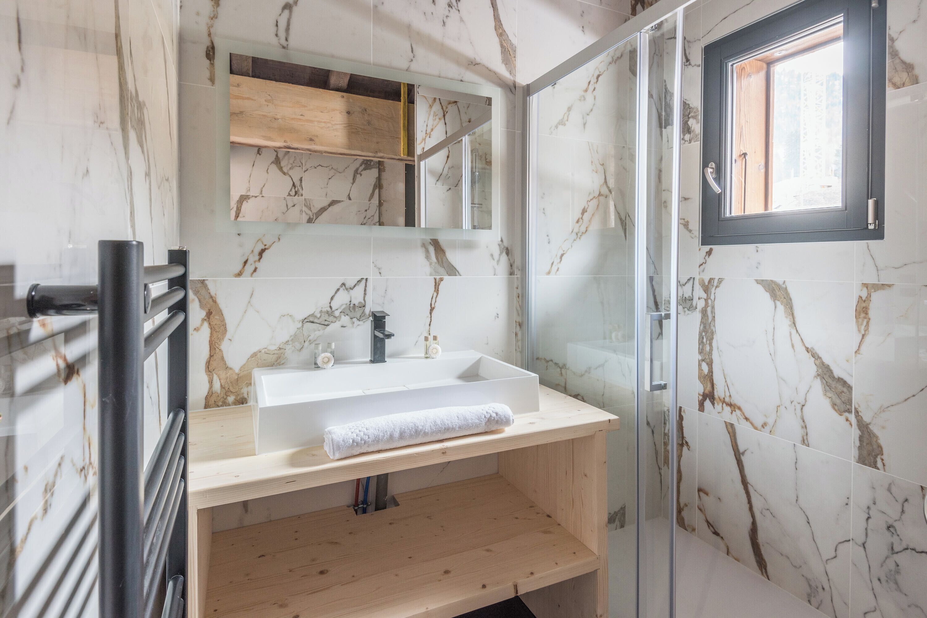 Un baño con salpicadero de mármol, tocador de madera, cabina de ducha de cristal y ventana.