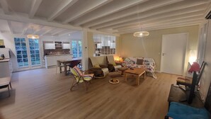 A spacious living area with a wooden floor, a sofa, a coffee table, and a dining table with chairs. There is a kitchen in the background with white cabinets and a window. - CAPBRETON. Villa ALAMEAT with garden for 6 people. Free Wifi. (CAPBRETON)