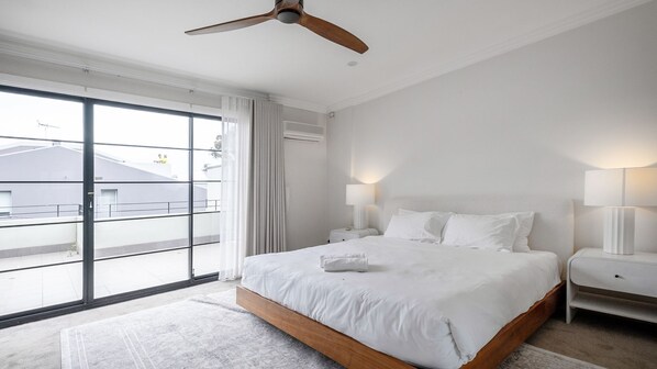 Un dormitorio moderno con una cama grande, dos mesitas de noche con lámparas, un ventilador de techo y una puerta corrediza de vidrio que da a un balcón.