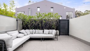 A modern outdoor seating area with a white sofa and a wicker chair, a black gate, and a building in the background. - The Euston Social in Alexandria (Alexandria)