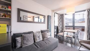 A modern hotel room with a grey sofa, a dining table, and a view of mountains through the window.
