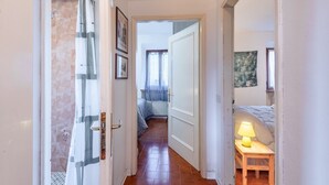 A hallway leading to a bedroom with a bed, a nightstand with a lamp, and a window with curtains.