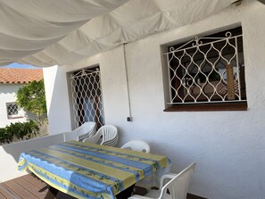 Ein überdachter Außenbereich mit Tisch und Stühlen, einer weißen Wand und einem Fenster mit einem Metallgitter.