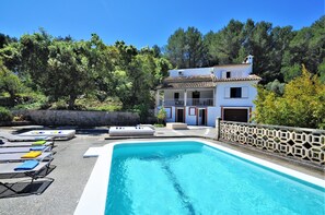 Una piscina con sillas de descanso, un edificio blanco con balcón y un paisaje montañoso cubierto de vegetación al fondo.