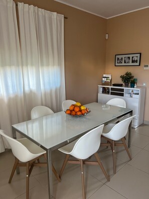 Een eethoek met een glazen tafel, witte stoelen, een fruitschaal, een wit kastje met een plant, en een raam met gordijnen.