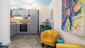 A compact kitchen with grey cabinets, a stove, and a dining area with a yellow armchair and teal stools.