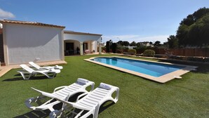Una piscina con césped artificial, sillas reclinables blancas y un edificio con techo de tejas.