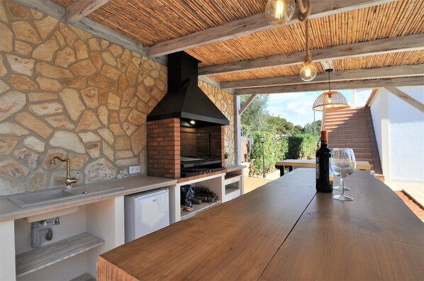 An outdoor kitchen with a stone wall, a brick oven, and a wooden table with a wine bottle and glass.