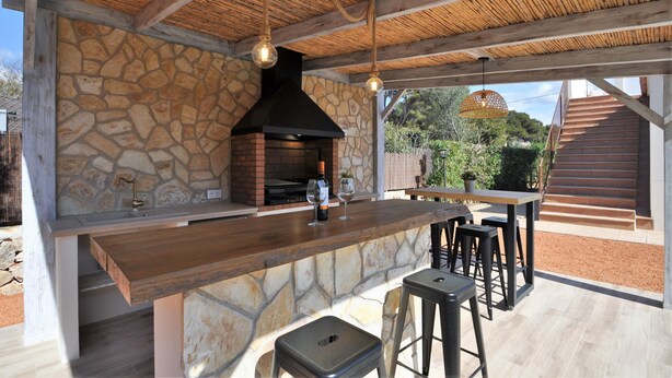 Cocina exterior revestida de piedra con una parrilla empotrada, barra de madera y taburetes.