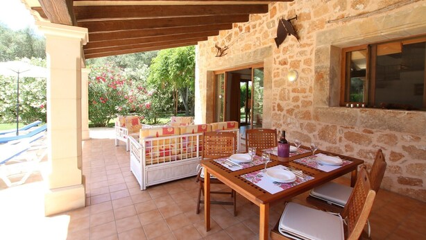 A covered outdoor dining area with stone walls, wooden furniture, and a view of the garden. - Country House 'Ca Na Barbara' with Private Pool, Wi-Fi, and Air Conditioning (Santanyí)