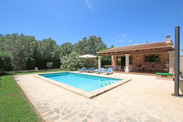 A swimming pool with lounge chairs, a covered patio with outdoor seating, and a stone building with a tiled roof.