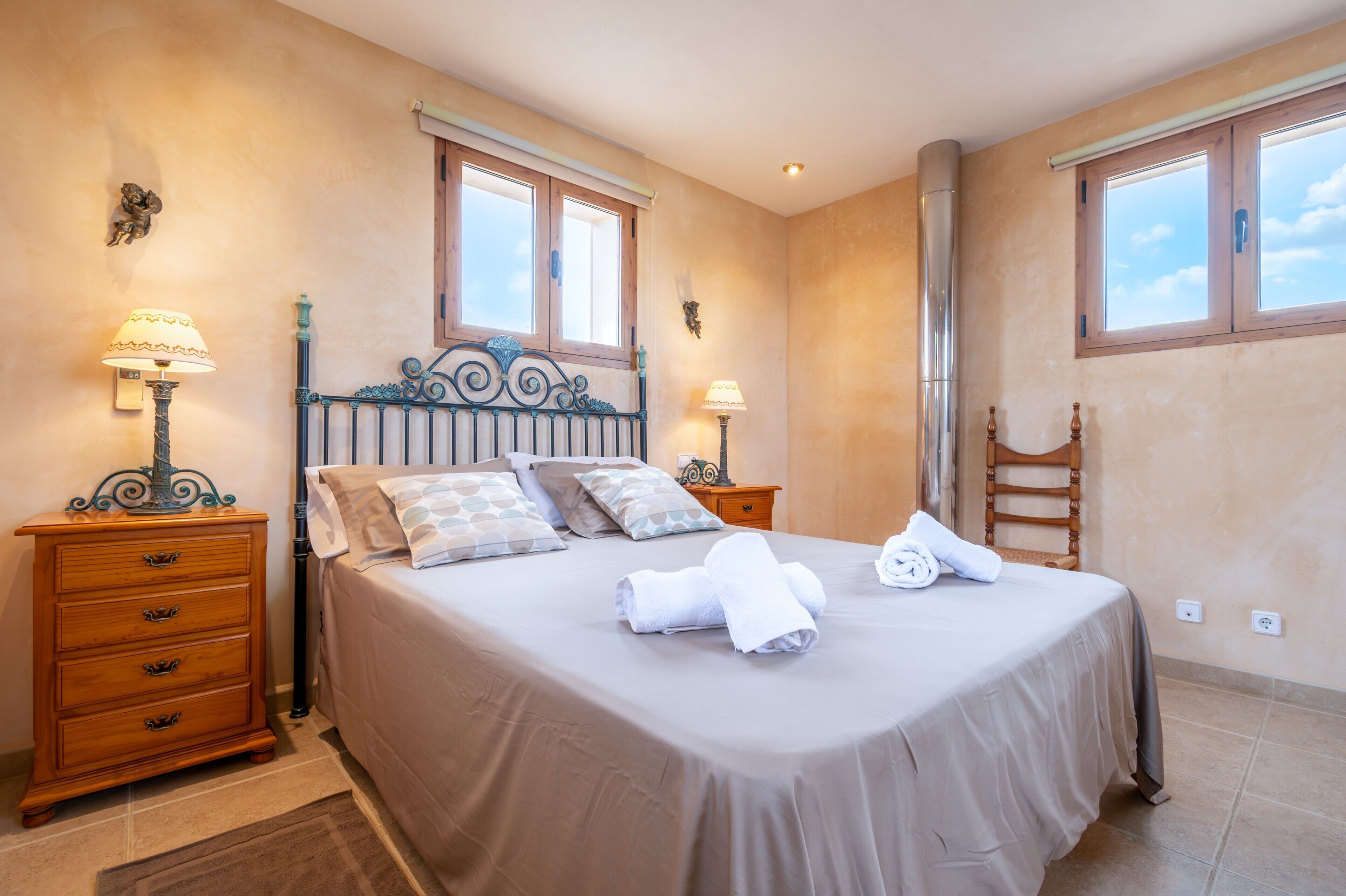 Una cama bien hecha con almohadas y toallas, un aparador de madera, dos lámparas de pared y una ventana con vistas.