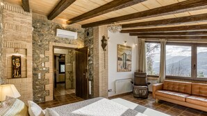 A room with a stone wall, a bed with patterned bedding, a leather sofa, and a view of mountains through large windows.