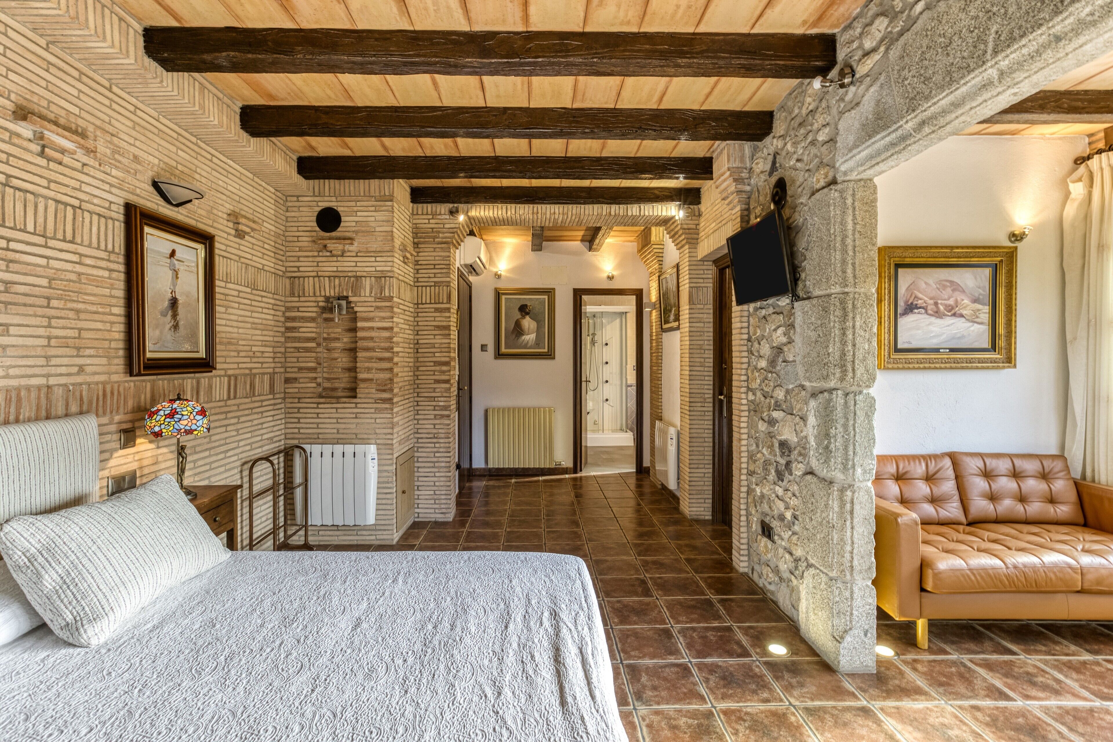 A bedroom with a bed, a nightstand, a lamp, a stone wall, a wooden ceiling, and a bathroom with a shower and toilet.