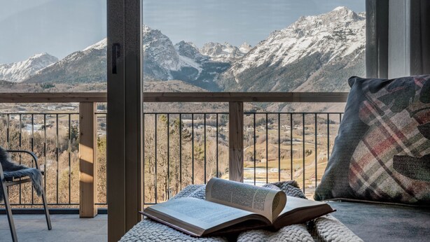 A balcony with a view of snow-capped mountains, a chair, a sofa with a plaid pillow, and an open book. - Apartment 'Rododendro' with Mountain View, Wi-Fi and Air Conditioning (Cavedago)