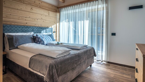 A bedroom with a bed, wooden ceiling beams, a window with curtains, and a bedside table.