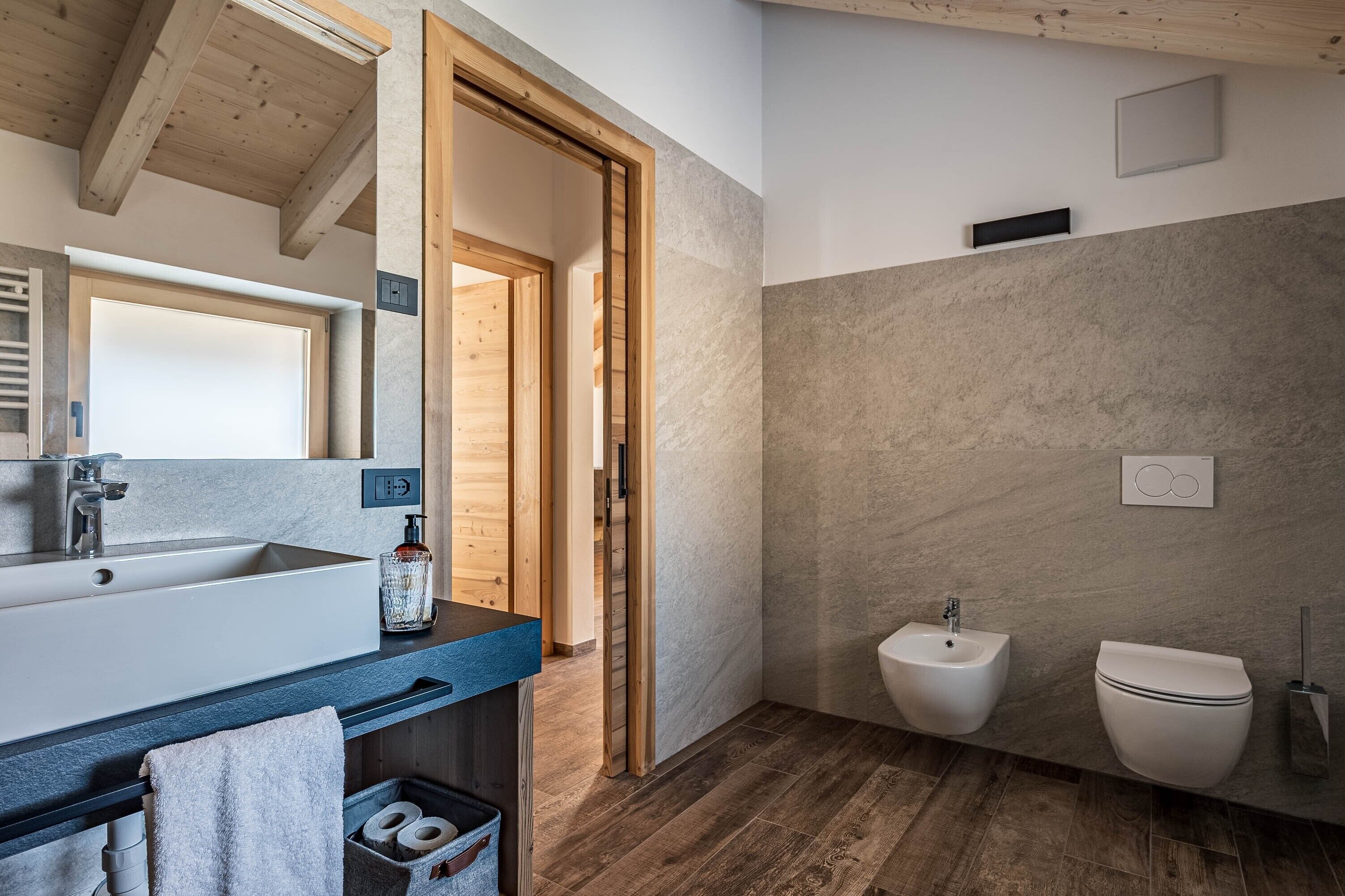 A modern bathroom with a large sink, bidet, and toilet, wooden beams, and a stone wall.