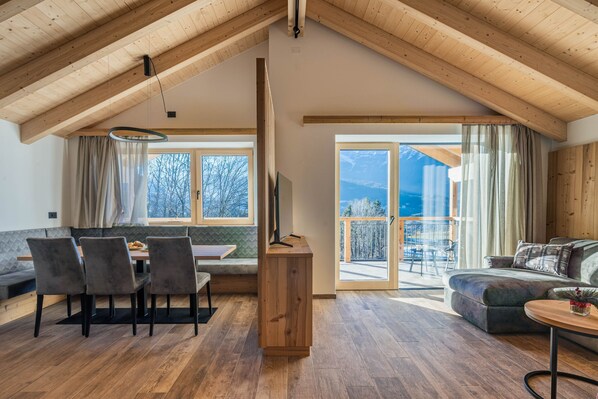 Un loft espacioso con techo alto, vigas de madera, una zona de comedor con un banco, un sofá y un balcón con vistas.