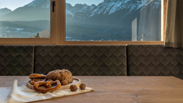 A table with pretzels and a loaf of bread, a window with a mountain view, and a cushioned bench.