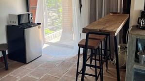Cocina compacta con refrigerador, microondas y cafetera. Hay una mesa de madera con dos taburetes y una ventana con cortinas translúcidas.