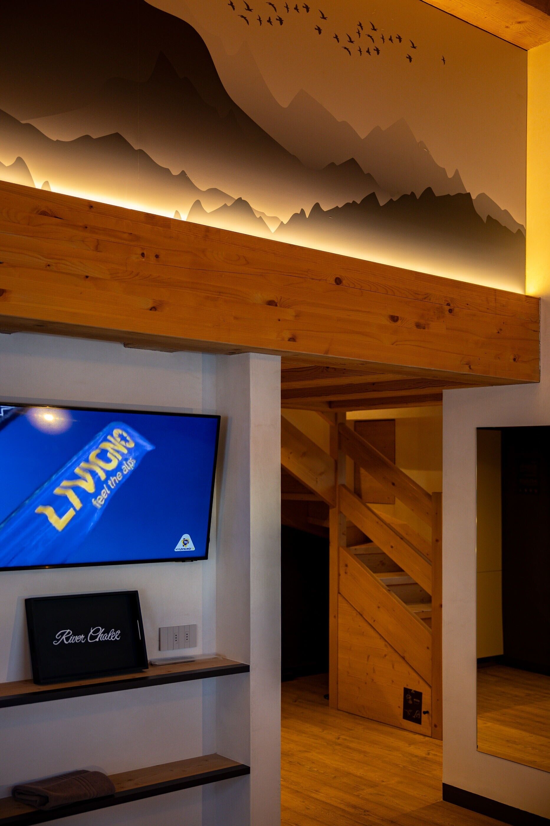 Una stanza con una TV che mostra 'LYRIC', un quadro incorniciato con l'etichetta 'River Chalet' e una scala in legno.