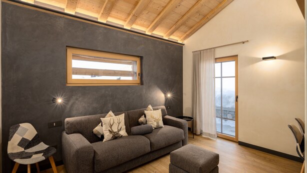 Ein modernes Wohnzimmer mit einer Couch, einem Hocker und einem Sessel. Ein Fenster lässt natürliches Licht herein, und eine Tür führt auf einen Balkon.