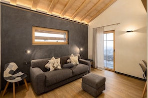 Un salon moderne avec un canapé, un pouf et un fauteuil. Une fenêtre laisse entrer la lumière naturelle, et une porte donne accès à un balcon.