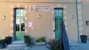 A building with two green doors, a sign reading 'RESTAURANT BAR A VINS CAVE,' and potted plants at the entrance. - Château des Gipières, exceptional apartment: Private Garden, Pool, View of Mont Ventoux (Montbrun-les-Bains)