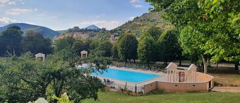 Une piscine entourée de chaises longues, bordée d’arbres et située au pied d’une colline où se dressent des bâtiments.