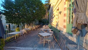 An outdoor patio with tables and chairs, a green fence, and a building with a brick facade. - Château des Gipières, exceptional apartment: Private Garden, Pool, View of Mont Ventoux (Montbrun-les-Bains)