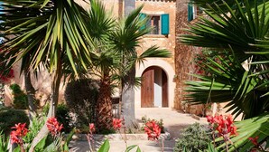A courtyard with a palm tree, red flowers, and a building with a wooden door and green shutters. - Country House 'Magdalena - Son Gaya' with Private Pool, Wi-Fi and Air Conditioning (Illes Balears)