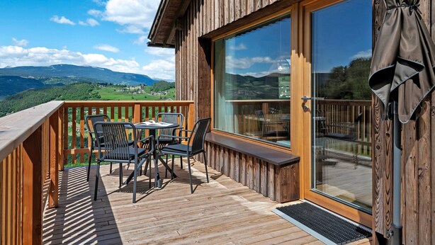 Eine Holzdirektlage mit Tisch und Stühlen, ein großes Fenster und Blick auf eine gebirgige Landschaft.