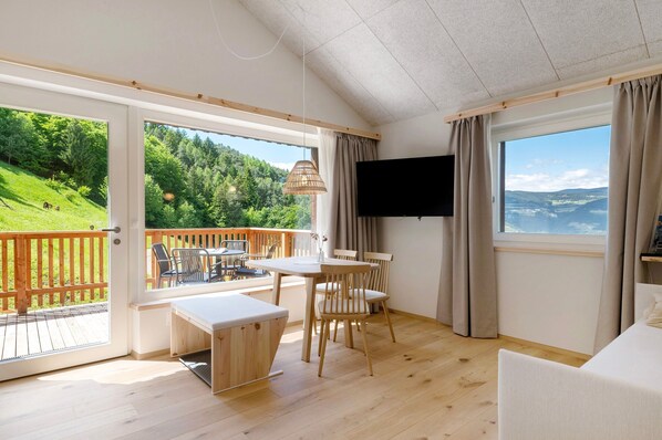 A room with a wooden floor, a table with chairs, a TV, and large windows with curtains. - Apartment 'Franz' with Mountain View, Private Terrace and Wi-Fi (Cornedo all'Isarco)