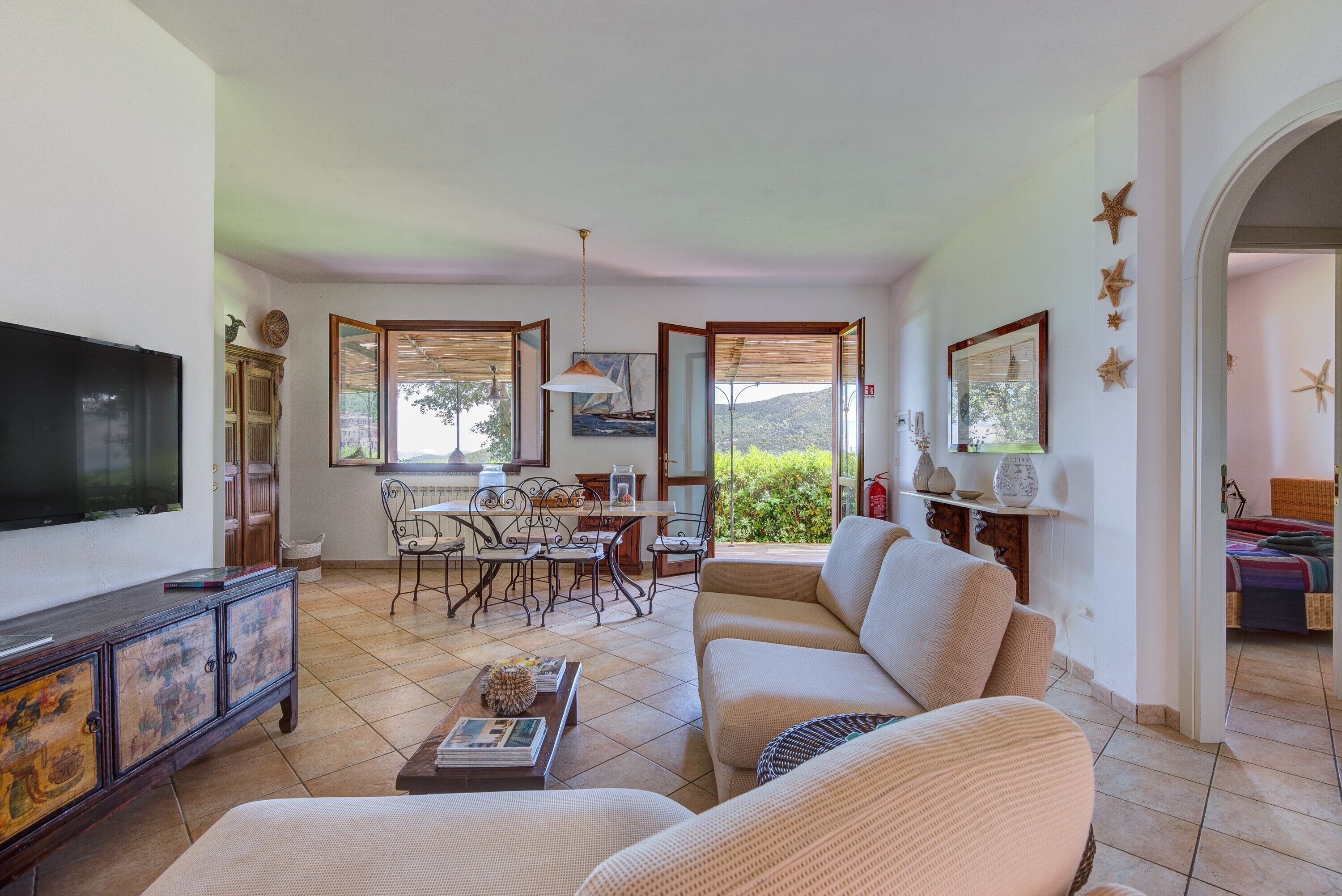 A living room with a TV, sofa, dining table, chairs, and a view of greenery outside.