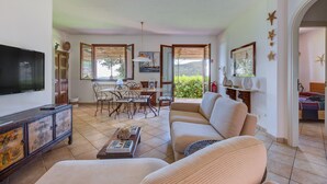 A living room with a TV, sofa, dining table, chairs, and a view of greenery outside. - Villa 'Martini' with Sea View, Private Terrace and Wi-Fi (Marciana)