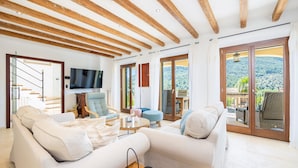 A spacious living area with a wooden ceiling, a flat-screen TV, and a view of a mountainous landscape through large sliding doors. - Villa 'Sa Papaiona' with Mountain Views, Private Pool and Wi-Fi (Valldemossa)