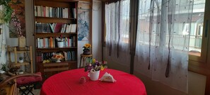Une pièce avec une table rouge, une bibliothèque et une fenêtre ornée de rideaux légers.