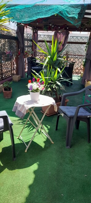 Un espace patio comprenant une table ronde, des chaises, des plantes en pot et une pergola surmontée d’un toit en treillis végétal.
