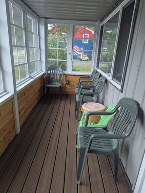 A covered porch with wooden flooring, plastic chairs, and a view of a red building with an American flag.