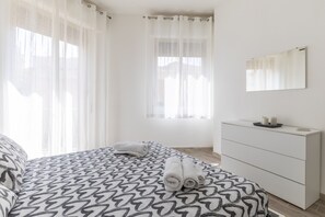 Une chambre à coucher avec une couvre-lit à motifs, une table de chevet avec des bougies et une grande fenêtre ornée de rideaux légers.