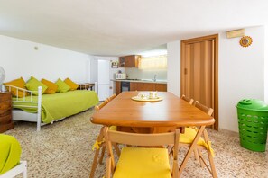 A compact living space with a kitchenette, a dining area with a wooden table and yellow chairs, a bed with green and yellow pillows, and a green trash bin. - Apartment 'Mi Casa Es Tu Casa - Cantina Giulia' with Private Terrace, Wi-Fi and Air Conditioning (Donnalucata)