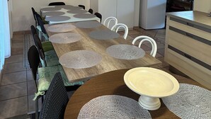 A dining area with a wooden table, white chairs, and a chandelier. There is a refrigerator and a coffee machine in the kitchen area.