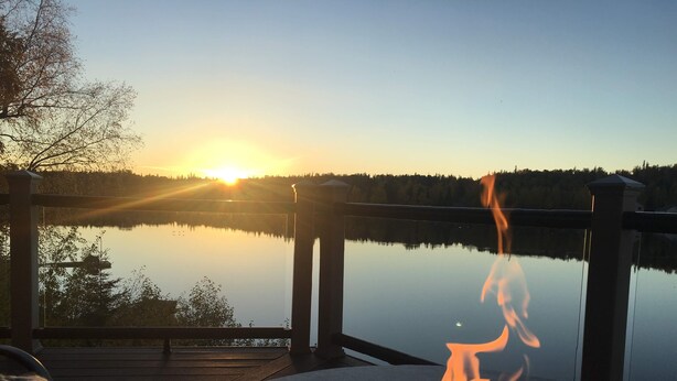 A serene lakeside deck with a fire pit and a glass table, overlooking a calm lake and a sunset.