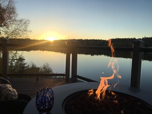 A serene lakeside deck with a fire pit and a glass table, overlooking a calm lake and a sunset. - Beautiful sunsets from the Lakeside Suite, deck on Longmere Lake, Soldotna AK (Soldotna)