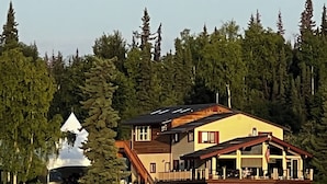 A large, multi-story house with a balcony, situated by a lake with a boat docked in the foreground.